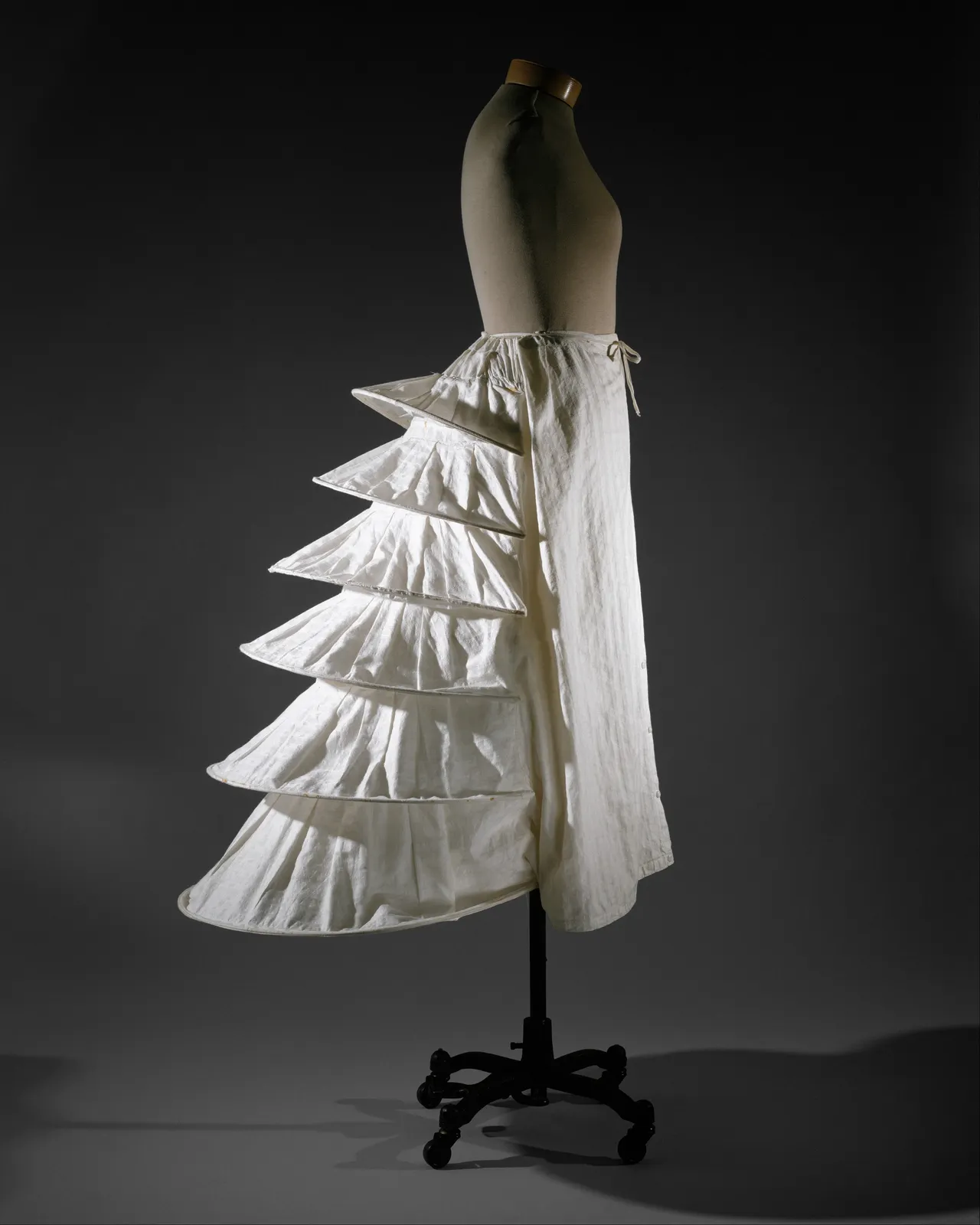 White cotton bustle seen from the side, flat front and stiffened tiers behind; British, about 1871; The Met 1985.27.4